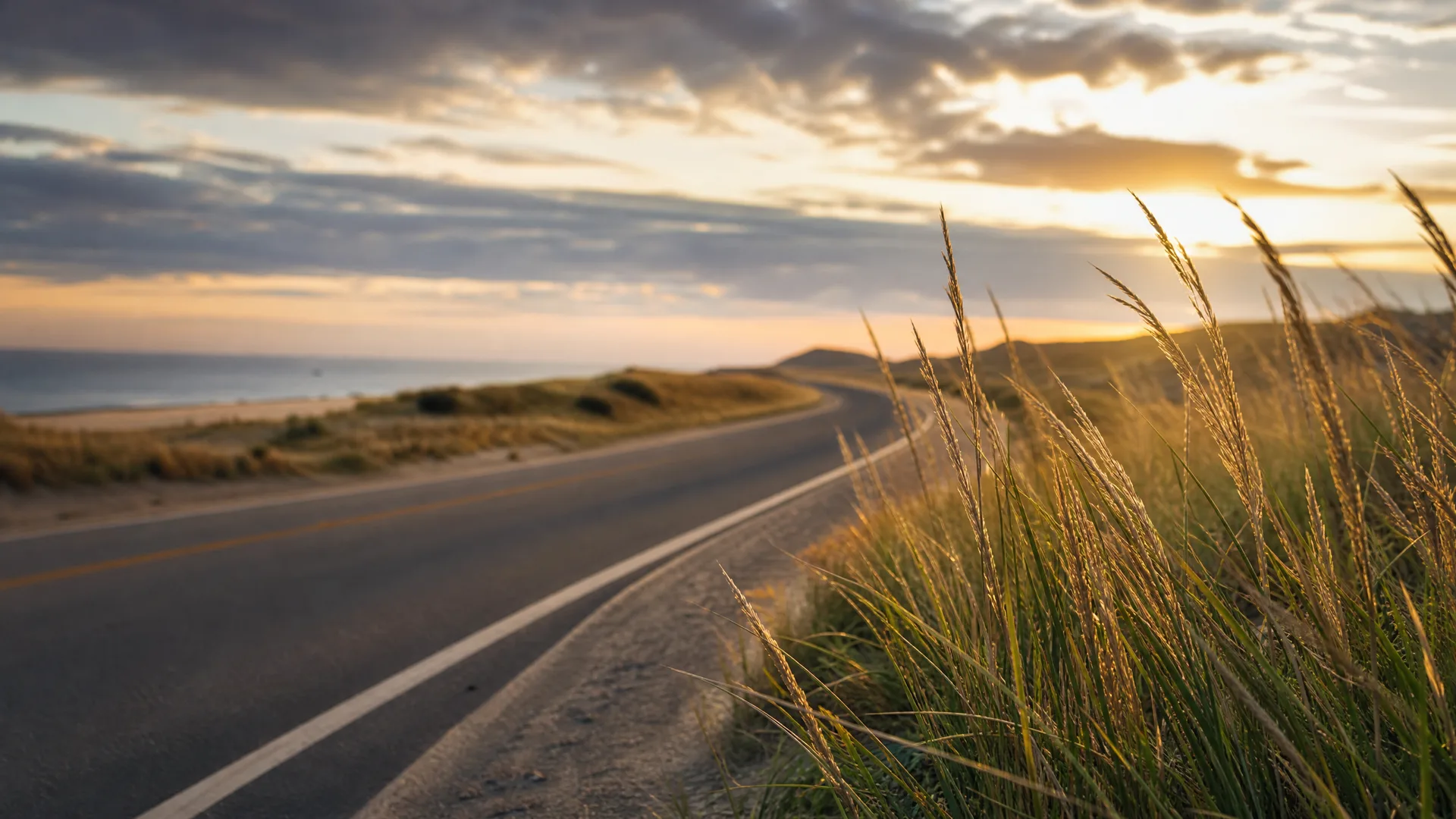 A two-lane paved road with a yellow center line and white edge line curves past grassy dunes toward low hills, with the ocean on the left and a cloudy sky lit by a low sun.